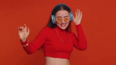 Happy pretty woman in headphones listening music and dancing disco fooling around having fun expressive gesticulating hands, relaxing on party. Caucasian girl isolated on red studio wall background