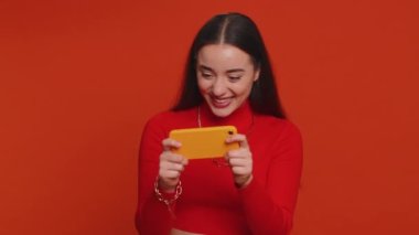 Worried funny addicted woman enthusiastically playing racing shooter video games on mobile phone. Young pretty girl using smartphone gadget app with drive simulator isolated on red studio background