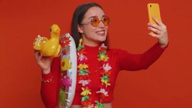 Happy young traveler blogger woman in modern sunglasses with swimming ring, luggage taking selfie on smartphone with subscribers record make video call online. Lovely tourist girl on red background
