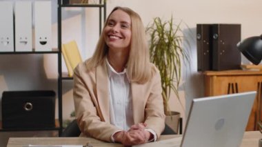 Modern ofis masasındaki gülümseyen sarışın genç iş kadınının portresi yüzünü kameraya çevir. Resmi takım elbiseli bir kadın. Güzel kadın. Bilgisayarda çalışırken uzak mesafeden çevrimiçi