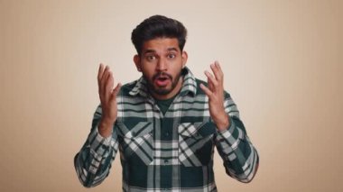 Oh my God, Wow. Excited amazed winner man raising hands in surprise looking at camera with big eyes, shocked by sudden victory. Sincere handsome indian guy isolated alone on beige studio background