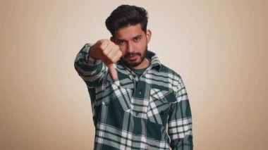 Dislike. Upset unhappy indian man in shirt showing thumbs down sign gesture, expressing discontent, disapproval, dissatisfied, dislike. Handsome young guy. Indoor studio shot on beige background