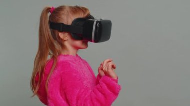 Rear view of child girl kid using headset helmet app to play simulation game. Watching virtual reality 3D 360 video. Little toddler female children in futuristic VR goggles on studio gray background