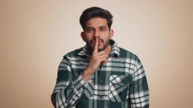 Shh be quiet please. Young indian man presses index finger to lips makes silence gesture sign do not tells secret, stop gossip. Handsome bearded hindu guy isolated alone on beige studio background