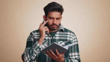 Thoughtful journalist indian man making notes, writing down thoughts with pen into notepad notebook diary, to do list, good idea. Handsome bearded hindu guy isolated alone on beige studio background