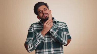 Indian bearded man touching sore cheek suffering from toothache cavities or gingivitis waiting for dentist appointment gums disease. Handsome hindu guy indoors studio shot isolated on beige background