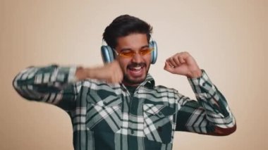 Happy indian man in headphones listening music and dancing disco fooling around having fun expressive gesticulating hands, relaxing on party. Hindu guy isolated alone on studio beige wall background