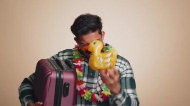 Traveler tourist indian man celebrating, dancing fooling with swimming inflatable duck toy. Concept summer vacation, journey trip to seaside. Bearded hindu guy in sunglasses on beige studio background