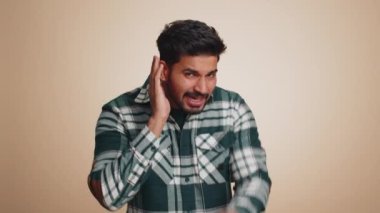I cant hear you. What. Young indian man trying hear, looking confused, frowning, keeping arm near ear for louder voice, asking to repeat, to hear information, deafness. Hindu guy on beige background