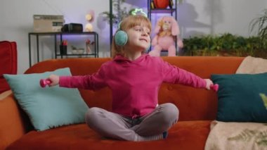 Young blonde child kid girl lifting dumbbells while doing exercises, enjoying training. Female teen toddler wearing headphones listening music at home in living room sits on sofa. Sport and fitness