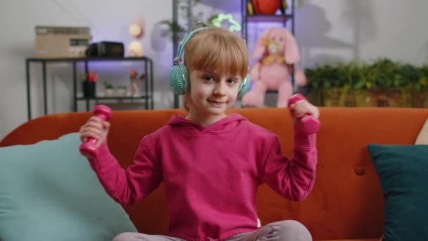 小女孩在做二头肌运动的同时举起哑铃 享受训练 在客厅里 女幼儿戴着耳机在家里听音乐 坐在沙发上 体育和健身 — 图库视频影像