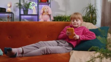 Young child kid girl texting share messages content on smartphone social media applications online, watching relax movie. Female teen toddler uses mobile phone at home in living room lying on sofa