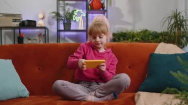 Worried funny child kid girl enthusiastically playing online racing or shooter video games on smartphone. Female teen toddler using smartphone gadget app with drive simulator at home sits on sofa
