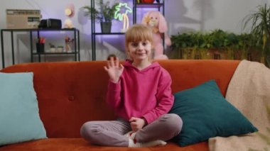 Young blonde child kid girl smiling friendly at camera and waving hands gesturing hello, hi, greeting or goodbye, welcoming with hospitable expression. Teen toddler at home in living room sits on sofa