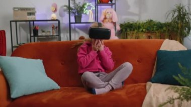 Genç sarışın çocuk modern sanal gerçeklik fütüristik teknoloji VR uygulama kulaklığı kullanarak 3D video oyunu simülasyonu yapıyor, film izliyor. Evde oturan genç bir kız çocuk kanepede oturuyor.