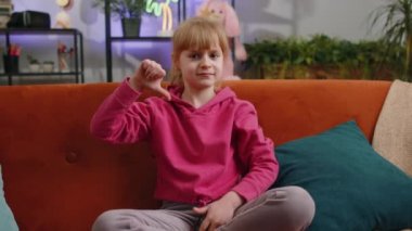 Dislike. Upset young blonde child kid girl showing thumbs down sign gesture, expressing discontent, disapproval, dissatisfied bad work, game fail. Female teen toddler at home in room sits on sofa