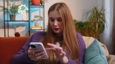 Woman sitting on sofa uses mobile phone smiles at home living room apartment. Portrait of young girl texting share messages content on smartphone social media applications online, watching relax movie