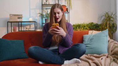 Happy woman use mobile smartphone typing browsing say Wow yes found out great big win good news celebrate lottery jackpot doing winner gesture. Portrait of young girl at home in room sitting on sofa