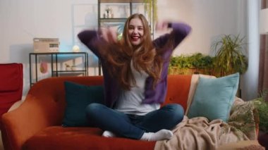 Happy young woman shouting, celebrating success, winning, goal achievement good news raising fists in gesture I did it, lottery luck at modern home apartment. Girl in living room sitting on couch
