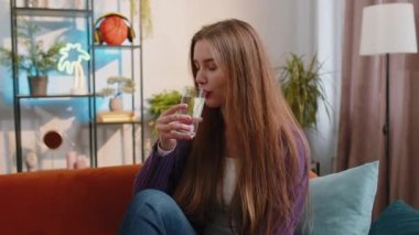 Thirsty young woman holding glass of natural aqua make sips drinking still water preventing dehydration sits at home living room. Girl with good life habits, healthy slimming, weight loss concept