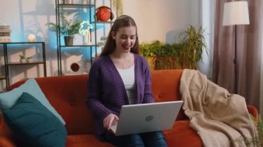 Young woman wearing headset, freelance worker, call center or support service operator helpline, having talk on laptop with client or colleague communication support at home room. Girl sitting on sofa