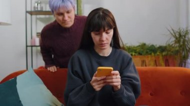 Woman freelancer is working on smartphone, shopping on e-commerce apps when her girl friend comes up behind and hugs her. Two young lesbian women family married couple at home living room. LGBT people