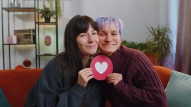 İki genç lezbiyen kadın aile çifti ya da kız arkadaşlar kalp şeklini kağıttan, sosyal medya işaretiyle kucaklaşıp evde sevgiyi gösteriyorlar. Homoseksüel kadın kucaklaşması. LGBT insan eşitliği. Neşeli, biseksüel