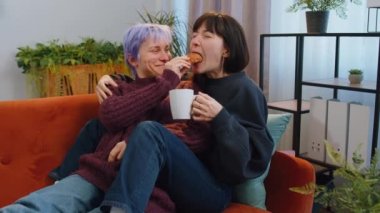 Two lesbian women family couple, girl friends drinking coffee or tea, eating croissants and talking at home morning living room. LGBT same-sex people. Enjoying happy breakfast together lying on sofa