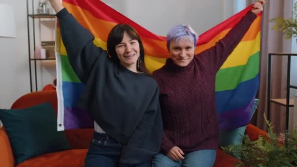 自宅のリビングルームでLgbtの人々のゲイの誇りフラグを手に持つ2つのレズビアン女性の家族のカップルや女の子の友人 ゲイバイセクシャルトランスジェンダーの社会運動 幸福の自由の概念は同性愛 — ストック動画