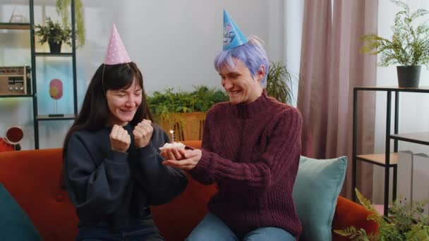 2人のレズビアン女性の家族カップルはお祝いの誕生日の帽子をかぶるカップケーキを保持している家庭のリビングルームでケーキの上に燃えるろうそくを吹くことを喜んでお祝いします 記念日パーティーを祝う女の子の友人 — ストック動画