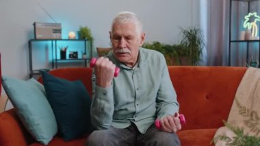 Büyükbaba evde halterli ağırlık kaldırma egzersizi yapıyor. Yaşlı adam kanepede oturma odasında antrenman yapıyor. Spor motivasyonu