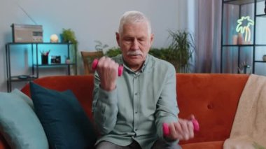 Olgun büyükbaba evde ağırlık kaldırma egzersizi yapıyor. Ev içinde. Yaşlı bir adam oturma odasında kanepede antrenman yapıyor. Spor motivasyonu