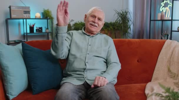 你好Hello 老爷爷在镜头前友好地笑着 挥挥手打招呼 打招呼或告别 在家里热情洋溢地欢迎 老年人坐在客厅的沙发上 — 图库视频影像