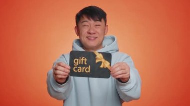 Yakışıklı Çinli adam hediye indirimi kuponu gösterip bayram indirimi kuponu satıyor. Alış veriş yapan Asyalı adam sürpriz kartı ile turuncu stüdyo arka planında tek başına.