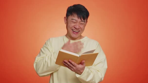 快乐的亚洲人阅读有趣有趣的童话故事书 业余爱好 知识智慧 一个英俊的中国人孤零零地躺在橙色的工作室墙上 — 图库视频影像