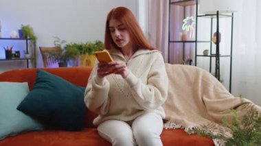 Hüzünlü genç kadın akıllı telefon daktilo kullanıyor, beklenmedik piyango sonuçlarını kaybediyor, kötü haber, servet kaybı, başarısızlık, teslim tarihi, virüs. Evdeki kanepede oturan kızıl saçlı kız
