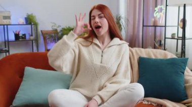 Tamam jest. Mutlu neşeli kadın kameraya olumlu bakıyor, işaret gibi olumlu, oturma odasında kanepede otururken güzel bir şeyi onaylıyor. Modern ev dairesindeki kızıl saçlı kız. Yaşam Tarzları