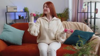 Kadın halterde ağırlık kaldırıyor, evde pazı kaldırıyor. Egzersiz, yağlı kalori yakma, antrenman gücü, ev içi spor aerobiği. Spor motivasyonu. Kanepede oturan kızıl saçlı kız.