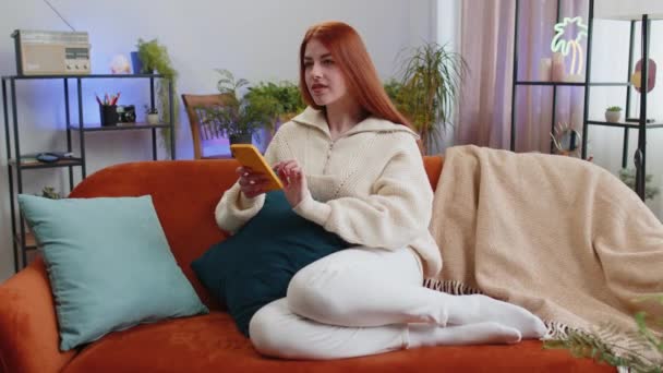 Рыжая Молодая Женщина Сидящая Диване Пользуется Мобильным Телефоном Улыбаясь Квартире — стоковое видео