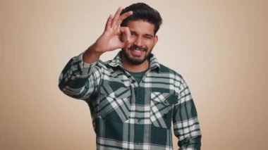 Tamam, tişörtlü neşeli mutlu Hintli adam kameraya olumlu bir geri bildirim hareketi göstererek olumlu bir işaret veriyor. Bej arka planda yalnız yakışıklı bir adam. İnsanlar samimi duygular