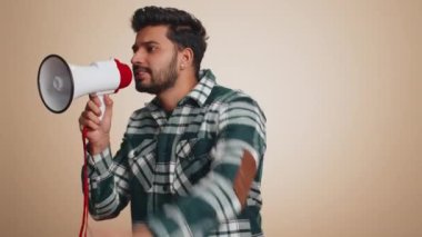 Hintli adam megafonla konuşuyor, haberleri duyuruyor, reklamları yüksek sesle duyuruyor, hoparlörü kullanarak konuşma yapıyor, boş yeri işaret ediyor. Kopyalama uzayı. Bej duvar arka planında yakışıklı bir adam.