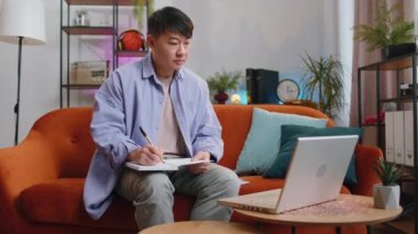 Asyalı yakışıklı adam web kamerasıyla konferans görüntülü görüşme yapıyor. Çevrimiçi öğretmen uzaktan sohbette e-öğreniyor. Çevrimiçi uzaktan eğitim. Çinli adam kanepeye oturmuş deftere not alıyor.