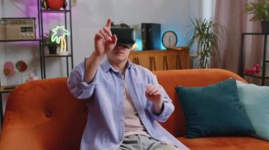 Asyalı bir adam sanal gerçeklik teknolojisi kullanıyor VR uygulama başlık simülasyonu 3D 360 video oyunu oynuyor ev dairesinde film seyrediyor. Gözlüklü adam kanepede oturuyor. Geçiş yakınlaştırma etkisi