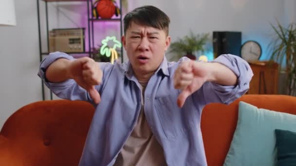 嫌いだ 親指をサインジェスチャーを示す動揺アジア人男性は 不満を表明し 屋内の現代の家庭のアパートで不満を抱えた悪い仕事 不満中国人男でリビングルームオンソファ — ストック動画