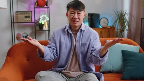 なぜだ アジア系の男性が怒りの表情で手を挙げているが 現代の家庭のアパートでのトラブルによる不信感を示す失敗の理由を尋ねる 中国人男でリビングルームでソファに座って — ストック動画