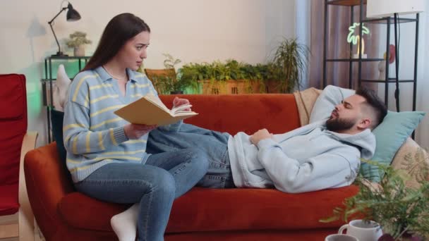 疲れた彼氏が眠っていびきをかいている間に本から物語を読む女 自宅で若い家族のカップル リビングルームでソファの上で一緒に夫の妻 趣味レジャー活動 — ストック動画
