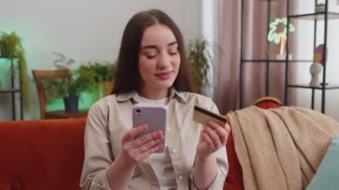 Woman using credit bank card and smartphone while transferring money, purchases online shopping cashless, order food delivery at home apartment indoors. Happy young girl sitting in room on couch