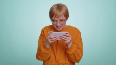 Endişeli komik bağımlı kadın cep telefonuyla yarış oyunları oynuyor. Mavi stüdyo arka planında izole edilmiş sürücü simülatörlü akıllı telefon uygulaması kullanan mutlu genç kız.