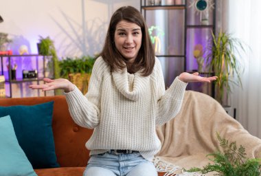 What. Why. Woman raising hands in indignant expression asking reason of failure demonstrating disbelief irritation by troubles at modern home apartment. Portrait of girl in living room sitting on sofa