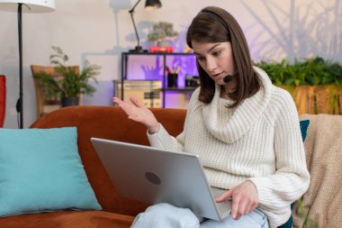 Kulaklık takan bir kadın, serbest çalışan, çağrı merkezi ya da destek operatörü yardım hattı, müşteri ya da iş arkadaşı ile iletişim desteği olan bir kadın, sınıf dairesinde el sallıyor. Kanepedeki kız